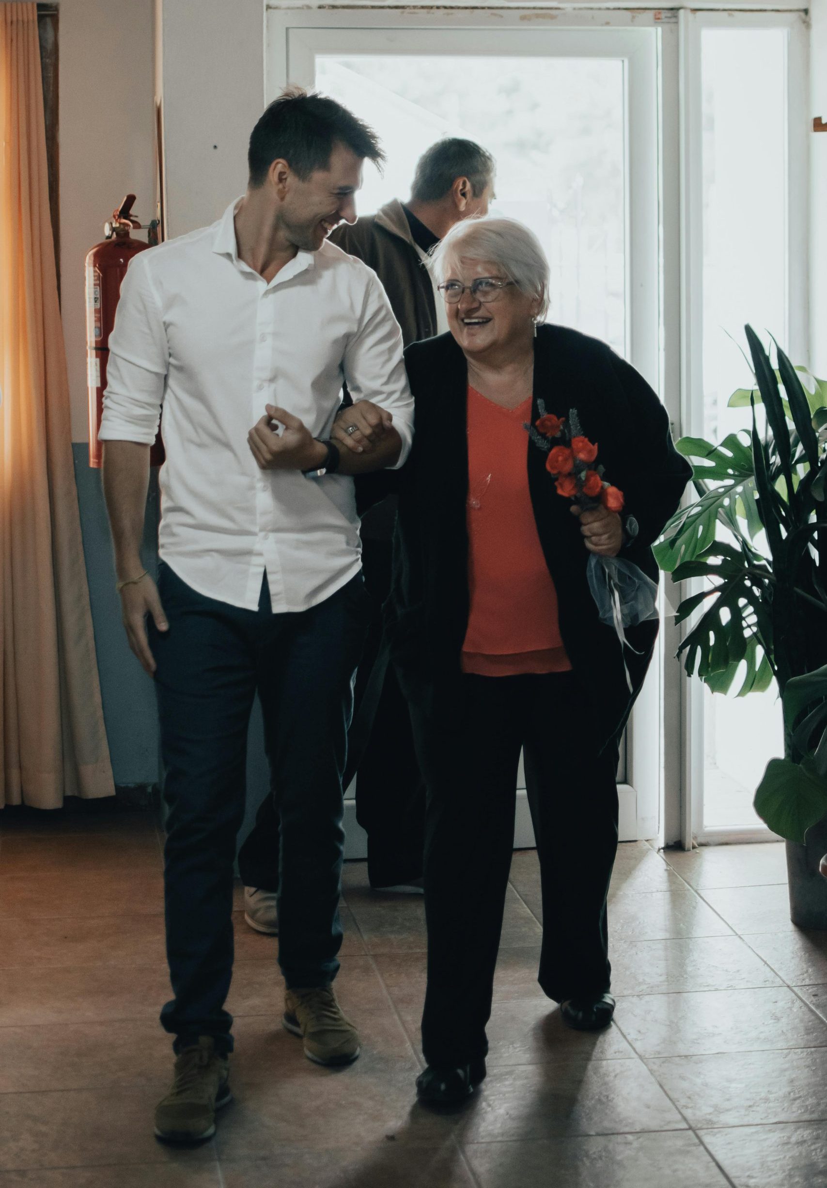 Senior woman arm in arm with a man, smiling and walking indoors with a bouquet of flowers.
